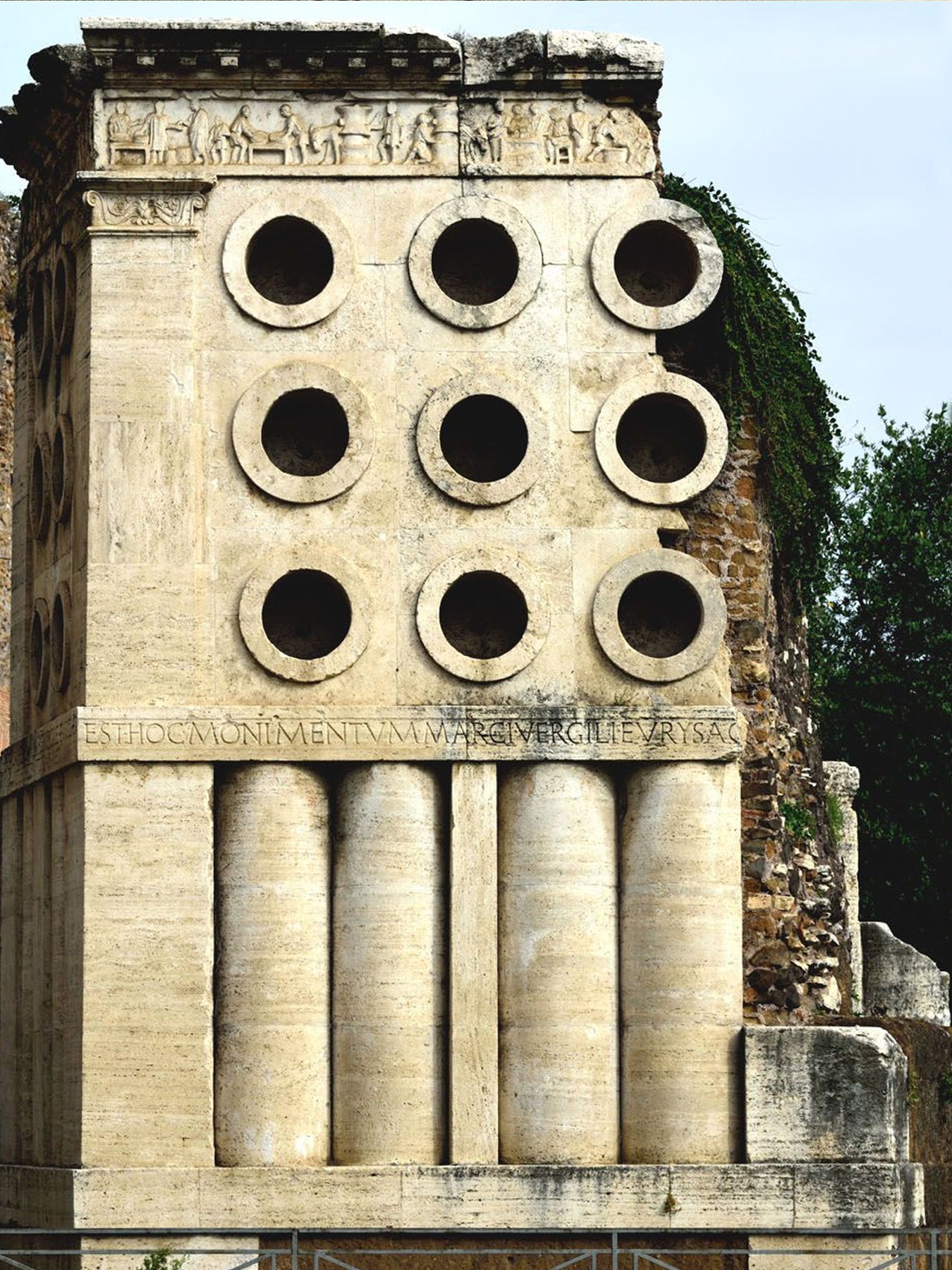 Ancient stone structure with circular holes and inscriptions, surrounded by greenery.