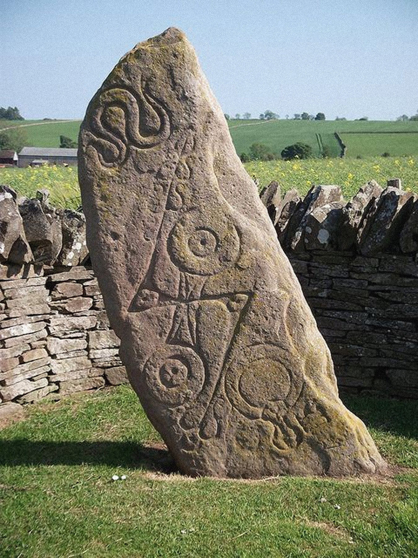 Decorative stone with Celtic designs in a field with a stone wall and greenery in the background