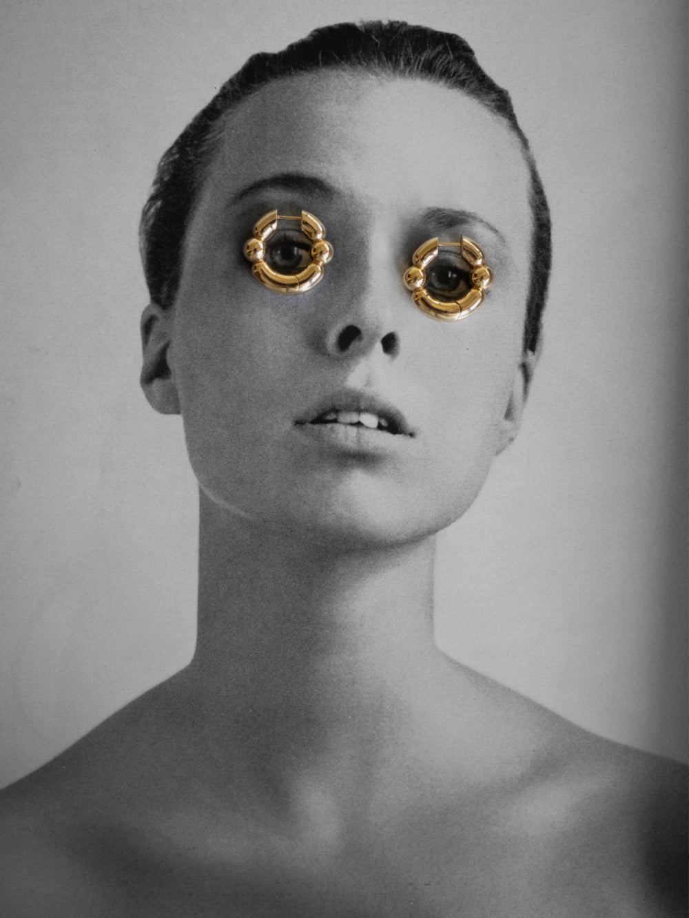 Black and white portrait of a person with gold rings over her eyes on a gray background