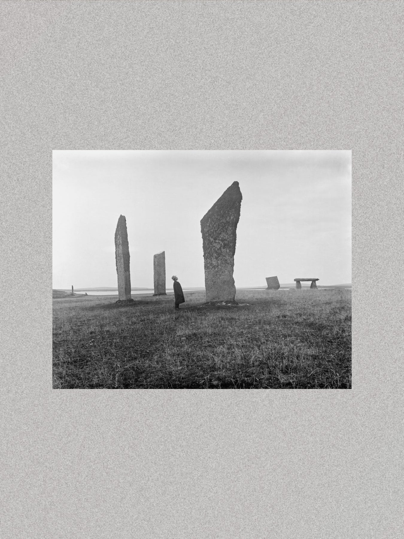 Black and white photograph of a person standing near stone monuments on a grassy field.