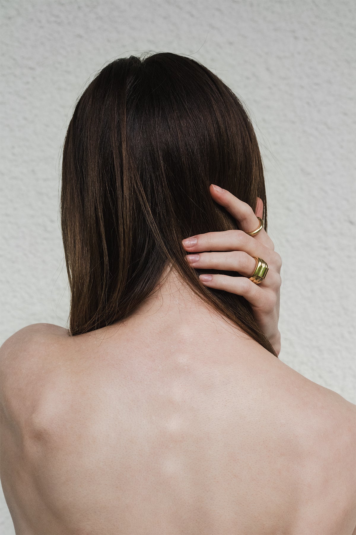 Person wearing two gold rings with long dark hair touching their neck against a neutral background
